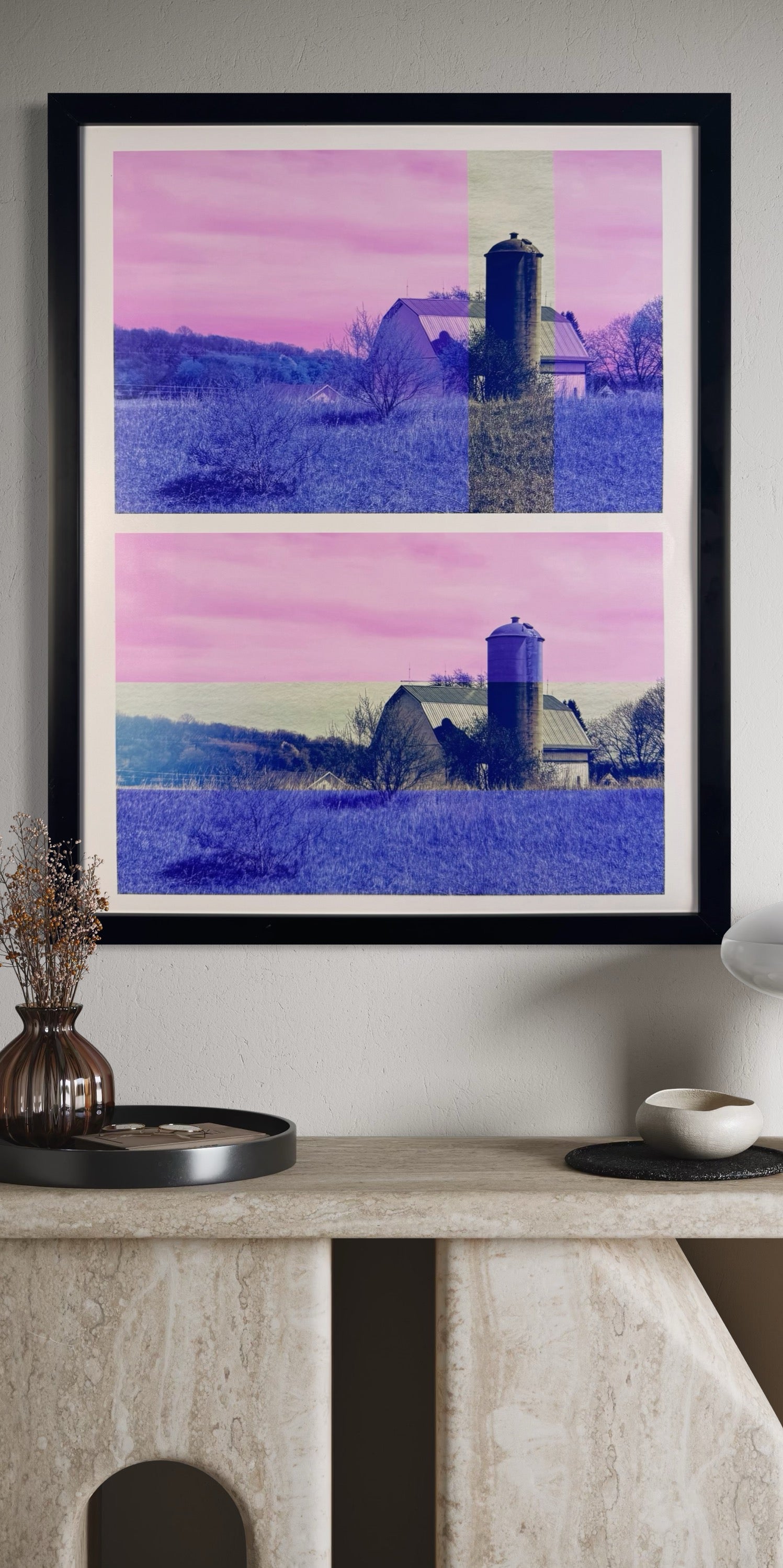 Framed artwork of a landscape with a silo on a white wall above a stone shelf.